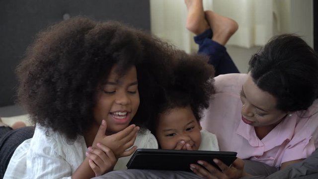 african american single mom with two daughter lying down on bed looking tablet computer together having fun stay at home . Happy black family with internet techmology .