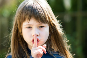 Portrait of pretty child girl with gray eyes and long hair holding point finger on her lips with hush sigh. Cute female kid on warm summer day outside.