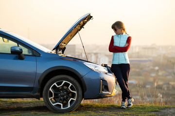 Young woman standing near broken down car with popped up hood having trouble with her vehicle....