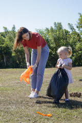 Young woman picking up plastic waste from the meadow with her toddler girl. Collecting garbage is educational activity to involve children in the protection of the environment.
