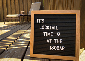 Wooden restaurant or pub sign with "locktail time" special at the "isobar" on a wood deck. Lounge chair in the background. Felt letter board with white letters. Covid-19 pandemic joke or meme.