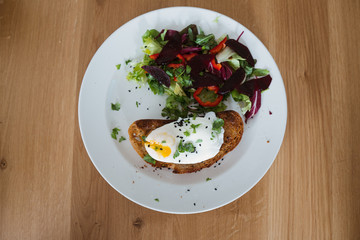 Poached egg on a piece of bread with green salad on a plate and on the wooden table. Healthy Breakfast