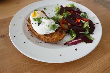 Poached egg on a piece of bread with green salad on a plate and on the wooden table. Healthy Breakfast