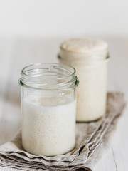 Two glass jars with wheat sourdough starters in different hydration levels. Starter 100 percent hydration in the background and starter with higher hydration in the foreground. Copy space. Vertical.