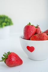 Fresh strawberries in white bowl with red heart on the white background with copyspace vertical photo