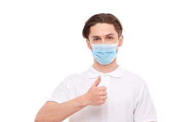 A man in a white T-shirt of European appearance in a medical mask, shows a thumb up. In the mask you are protected from covid 19 viruses.