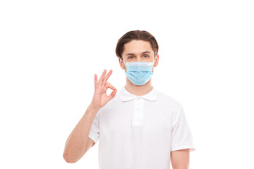 A man in a white T-shirt of European appearance in a medical mask, shows a thumb up. In the mask you are protected from covid 19 viruses.