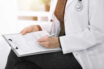 Unknown woman-doctor writing something at clipboard while sitting in sunny clinic, close-up. Therapist wearing green blouse at work is filling up medication history record. Medicine concept