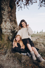 Stylish young family in the autumn mountains. Mom and daughter are sitting under a large old tree and hugging against the background of the forest and mountain peaks at sunset.