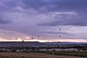 A unique sight. Many colorful balloons rise in the sky over the plain of Cappadocia. The sun rises and turns dark clouds pink. In the distance are the silhouettes of mountains.