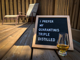Wooden restaurant or pub sign with "quarantini" and "triple distilled" placed on a wood deck. Lounge chair in the background. Felt letter board with white letters. Covid-19 pandemic joke or meme.