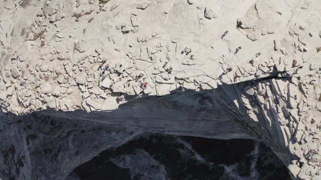 Aerial view of the flat top of Half Dome cliff brightly lit with the sun, with several tourists on it, walking near the edge. The steep stone slope of the mountain abruptly goes down. Yosemite. 4K
