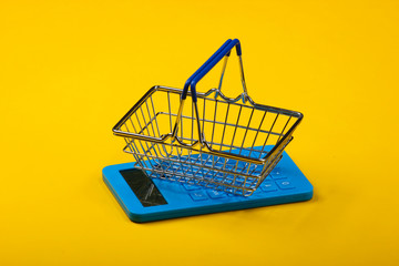 Calculator and shopping basket on yellow background. Top view