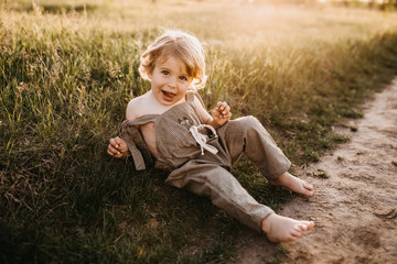 Little blonde boy wearing vintage jumpsuit, sitting in a field at sunset.