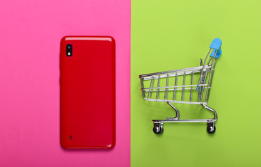 Mini shopping trolley with smartphone on pink green background. Top view