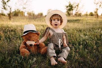 Little blonde boy wearing vintage jumpsuit and a straw hat, sitting in a field at sunset with a...