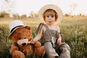 Little blonde boy wearing vintage jumpsuit and a straw hat, sitting in a field at sunset with a...