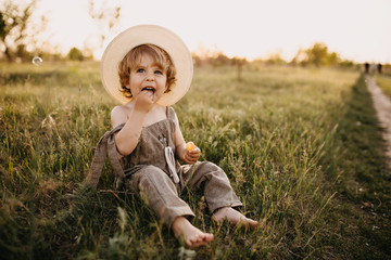Little blonde boy wearing vintage jumpsuit and a straw hat, sitting in a field at sunset.