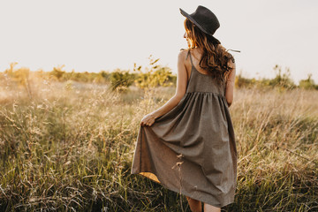 Young brunette woman with long wavy hair, wearing vintage sundress and a hat, walking in a golden field, at the sunset. - obrazy, fototapety, plakaty
