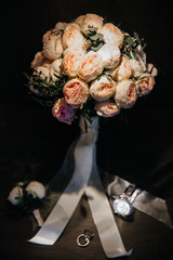 wedding bouquet with peony roses in light shades on a dark background and with a cozy light source on the bouquet. at the foot of the bouquet wedding accessories in the form of a buttonhole, rings and