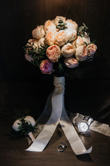 wedding bouquet with peony roses in light shades on a dark background and with a cozy light source on the bouquet. at the foot of the bouquet wedding accessories in the form of a buttonhole, rings and