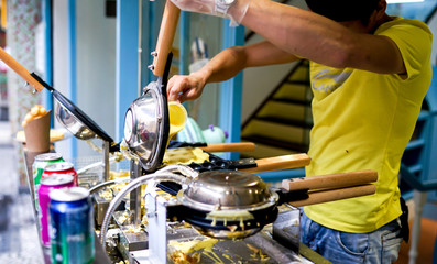 An Asian man in a yellow uniform is cooking french waffles on the streets, street food in Asia, the process of making sweet dessert