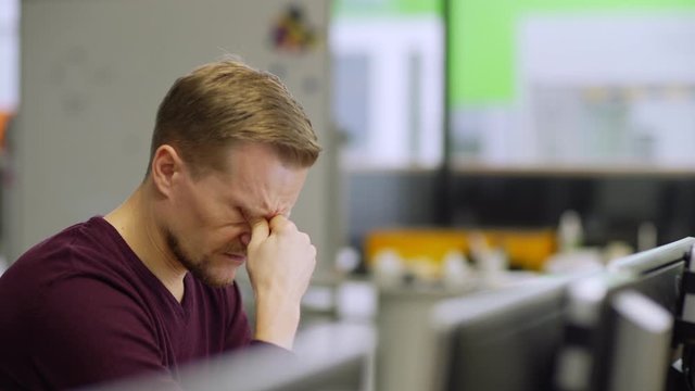 Tracking side view shot of middle aged engineer working on computer and rubbing tired eyes sitting at desk in laboratory