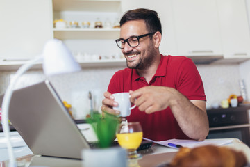 Happy young man, wearing glasses and smiling, as he works on his laptop to get all his business done early in the morning.