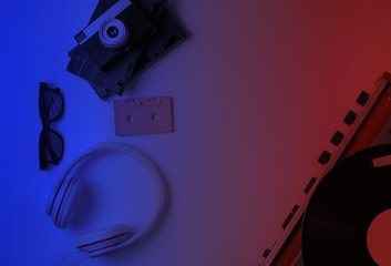 Flat lay retro 80s pop culture objects. Vinyl player, headphones, audio cassette, video tapes, film camera. Red blue neon gradient light