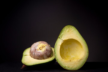Two halves of a large avocado fruit with a huge seed and ripe vibrant yellow green pulp. Studio low key food still life against a dark grey background.