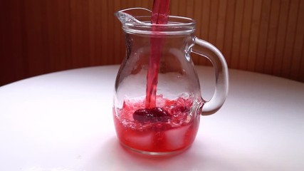 Raspberry lemonade is poured into a glass jug with pieces of ice and raspberries in it, standing on a white table, slow motion