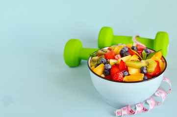 Healthy food. Bowl of vegetarian fresh fruit salad made of different fruits, dumbbells, measuring tape: banana, kiwi, apple, orange, strawberries, blueberries on blue background. The concept of diet