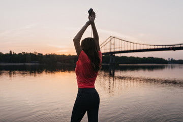 Fototapeta premium Enchanting shapely girl standing with hands up and looking at river. Outdoor photo from back of amazing female model relaxing after training near lake.