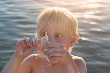 Child stares at shell. Funny fair-haired boy playing with shell on sea background