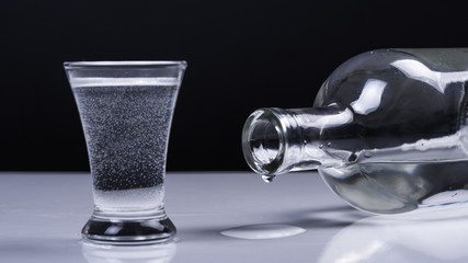 A glass of vodka and a reclining bottle with a drop on a white table and on a black background