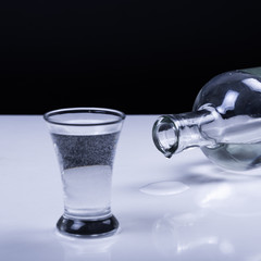 A glass of vodka and a reclining bottle with a drop on a white table and on a black background