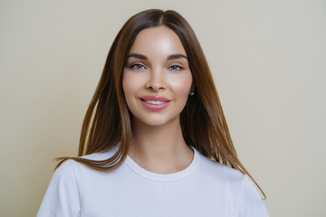 Fototapeta premium Headshot of beautiful dark haired young European woman looks with tender expression, has friendly attitude, talks casually with friends, wears casual white t shirt, isolated over beige background