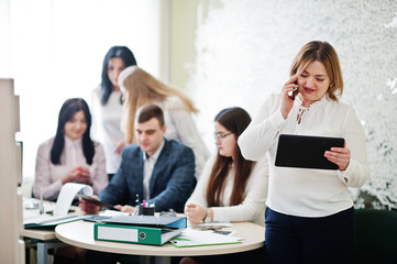 Portrait of caucasian woman in white blouse hold tablet and speak on phone against business people group of bank workers.