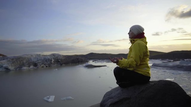 A girl in a bright yellow jacket meditates in a yoga pose on a stone in front of a huge glacier. Relaxation overlooking the mountains and cliffs