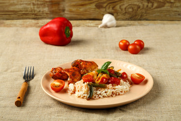Baked chicken wings in tomato sauce with rice and vegetables on a plate on a linen tablecloth next to vegetables for decoration.