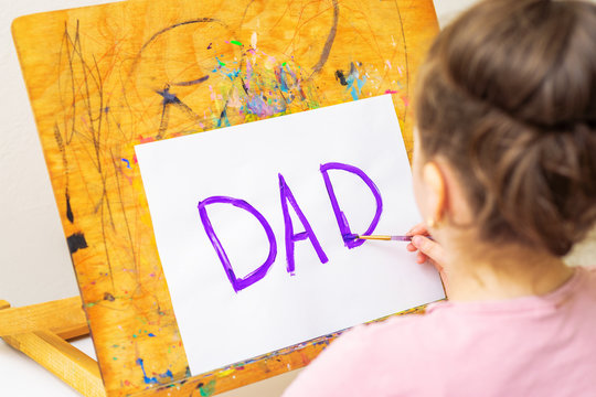 Child is drawing purple word Dad greeting card on white paper on easel. Happy Father's Day concept.