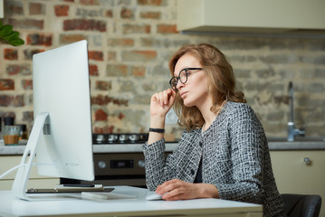A pretty woman in glasses works remotely on a desktop computer in her studio. A female boss searching for information at a video conference at home. A female professor preparing for an online lecture.