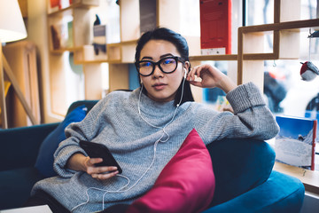 Obraz premium Pensive asian female millennial listening playlist song using app on smartphone and earphones, serious chinese woman 20s sitting at cozy sofa in home interior enjoying leisure and player via accessory