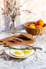 Breakfast. Fried eggs of two eggs and green feathers of onions. Sausage and bread on a wooden board. Sprigs of blossoming apple tree in a crystal vase. Apples and oranges in a wicker basket.