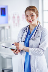 Young woman doctor is standing with board with clipboard smiling in hospital office