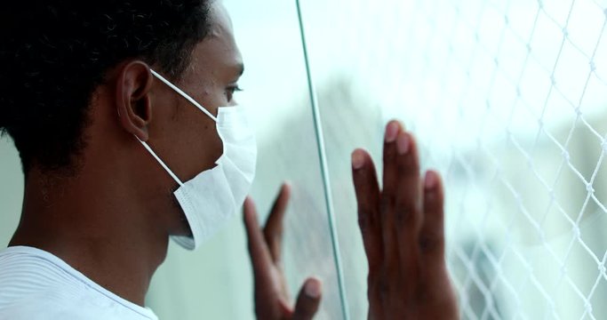 African american man during quarantine wearing covid virus mask indoors leaning on window looking outside