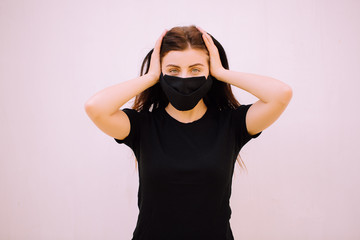 A young brunette girl in a black medical mask and black t-short covers her ears with her hands isolated on white background. Closed up photo. Free space for logo.