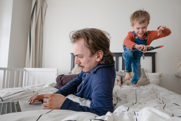 Man trying to work via laptop at home during quarantine with toddler boy jumping on bed . Family life during self isolation. Stay home. Work from home. home office