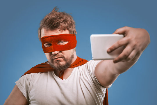 Super hero man takes selfie by mobile phones. Confident man in a super hero costume in a red mask and cloak poses on faded denim blue background making selfie photos on smartphone