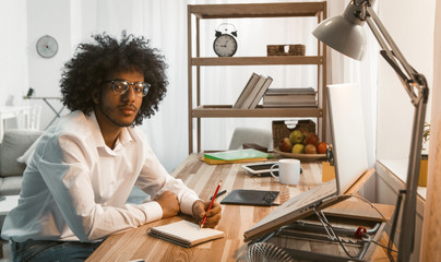 Creative man writes in Note Pad working laptop at Home work place. Arabic business or student in eyeglasses looking at camera. Freelance concept. Work from home concept